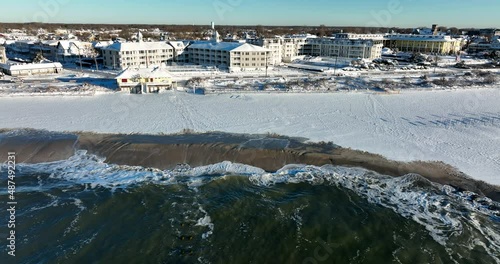 New Jersey beach town in winter snow. Sand covered in white snowfall. Waves crash at oceanfront hotel on seaside view. Truck shot aerial.