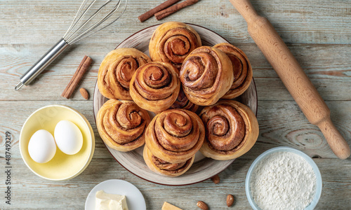 There is a plate with homemade fresh delicious cinnamon rolls, ingredients for their preparation and kitchen tools on the wooden table . Top view
