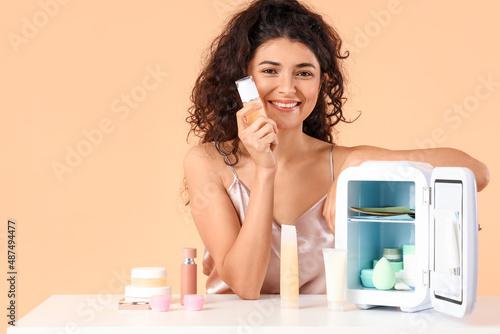 Young woman near table with opened refrigerator and cosmetic products on color background