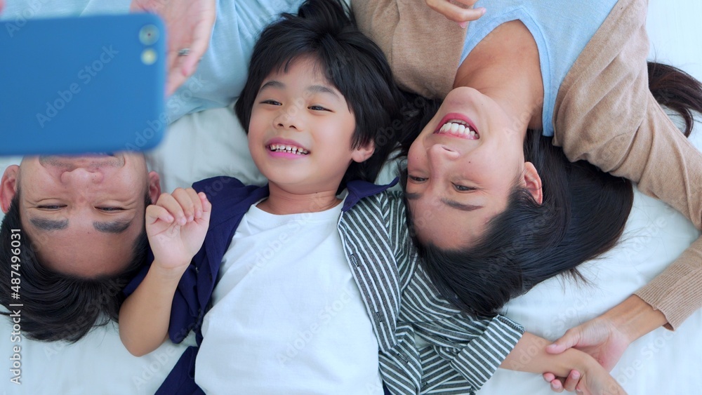 Happy family time, Taking selfie mobile phone of Asian parents dad, mom and son laying together on bed in bedroom with happy, laughing smiles. Weekend parenting with love good time relaxing at home. Happy family time, Taking selfie mobile phone of Asian parents dad, mom and son laying together on bed in bedroom with happy, laughing smiles. Weekend parenting with love good time relaxing at home.