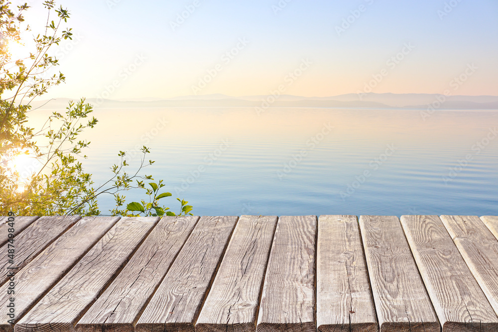 An empty wooden deck or table against the backdrop of a calm morning lake at dawn in pastel colors. Product display frame.