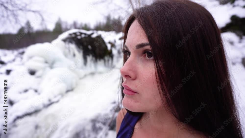 A beautiful girl in a blue dress at a frozen waterfall looks into the distance. Woman in the winter forest near the river between the rocks. Portrait of a brunette in a snowy forest A beautiful girl in a blue dress at a frozen waterfall looks into the distance. Woman in the winter forest near the river between the rocks. Portrait of a brunette in a snowy forest