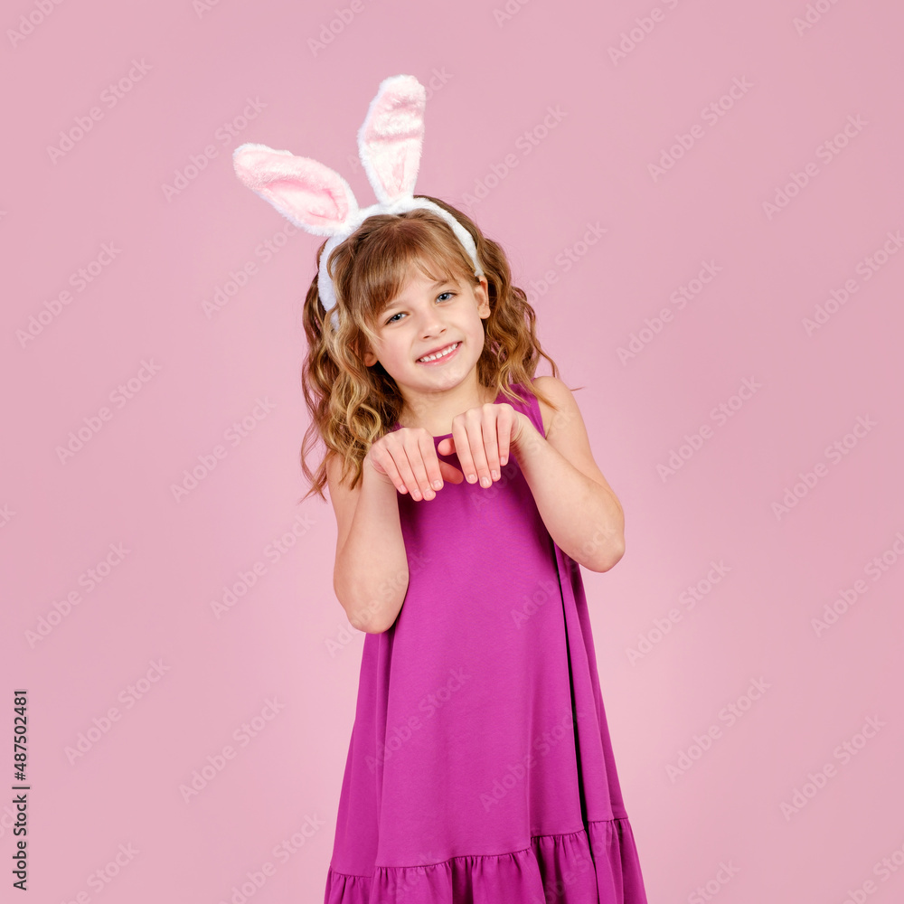 Adorable funny little kid with curly blond hair in dress and bunny ears, smiling and showing paws gesture while imitating rabbit during Easter party in pink studio