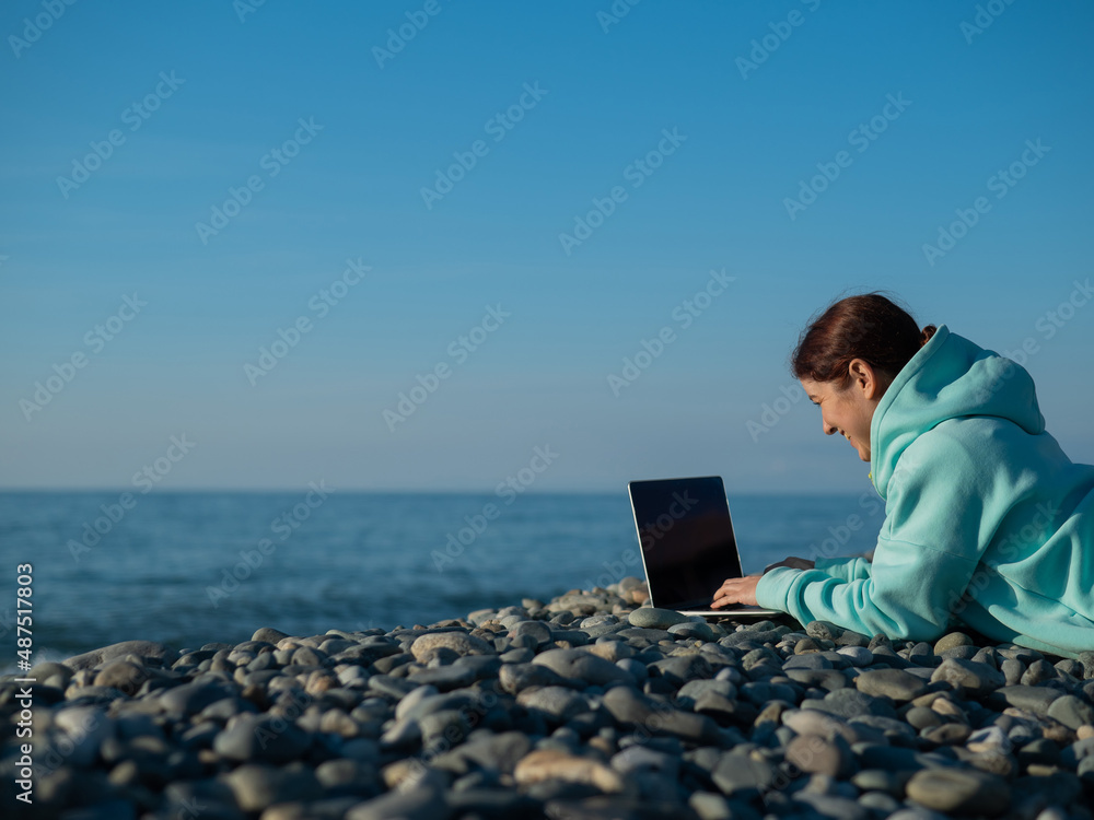 Caucasian woman lying on her stomach on a pebble beach and typing on a laptop. Remote work freelancer.