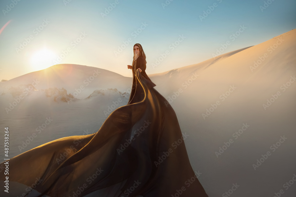 Mystery arabic woman in black long dress stands in desert long train silk fabric fly flytter in wind motion. clothes gold accessories hide face. Oriental fashion model. Sand dunes background sunset. - obrazy, fototapety, plakaty 