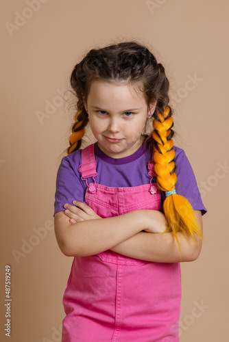 Offended small girl having yellow kanekalon braids looking at camera askance with folded arms wearing pink jumpsuit and purple t-shirt on beige background.