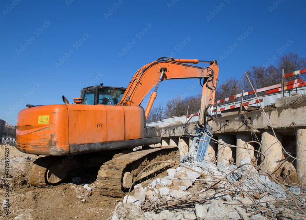 orange excavator with an industrial hydraulic hammer is working on a construction site. Pieces of concrete and metal fittings. orange excavator with an industrial hydraulic hammer is working on a construction site. Pieces of concrete and metal fittings.