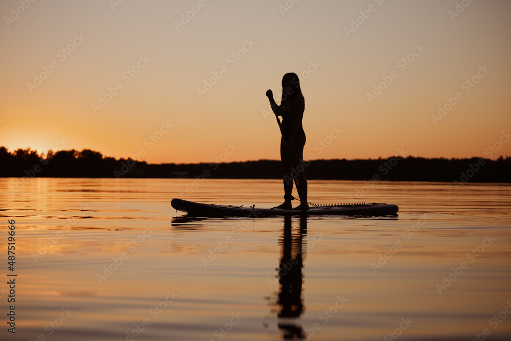 Caucasian fit woman silhouette sup boarding having paddle in hands on bay with reflection on water at sunset time in summer. Active lifestyle. Exercising. 