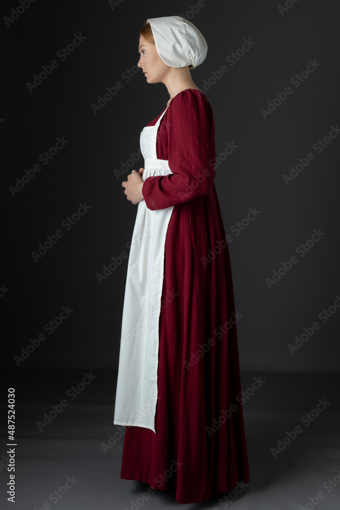 A Regency maid servant wearing a red linen dress with an apron and a cap against a studio backdrop A Regency maid servant wearing a red linen dress with an apron and a cap against a studio backdrop