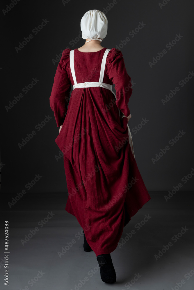 A Regency maid servant wearing a red linen dress with an apron and a cap against a studio backdrop A Regency maid servant wearing a red linen dress with an apron and a cap against a studio backdrop