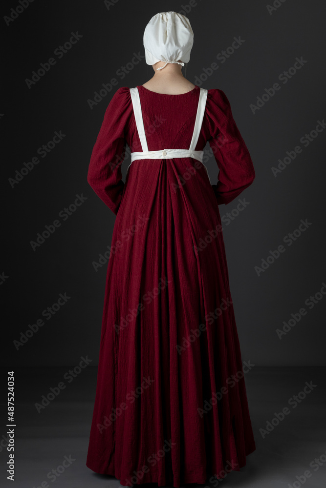 A Regency maid servant wearing a red linen dress with an apron and a cap against a studio backdrop A Regency maid servant wearing a red linen dress with an apron and a cap against a studio backdrop