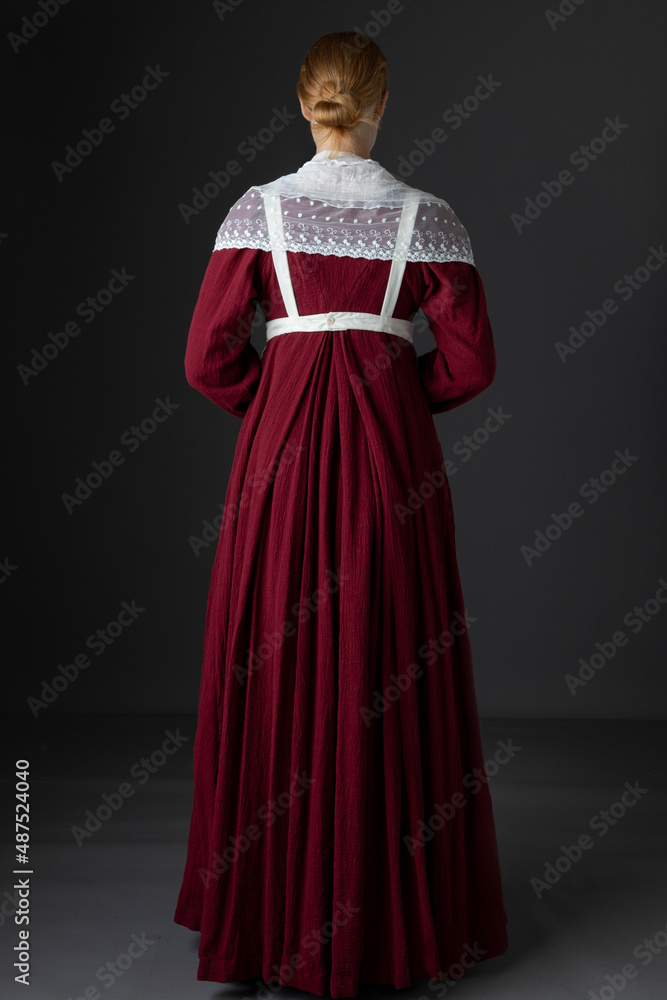 A Regency maid servant or working woman wearing a red linen dress with an apron and a lace modesty shawl against a studio backdrop