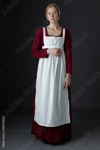 A Regency maid servant wearing a red linen dress with an apron and a cap against a studio backdrop