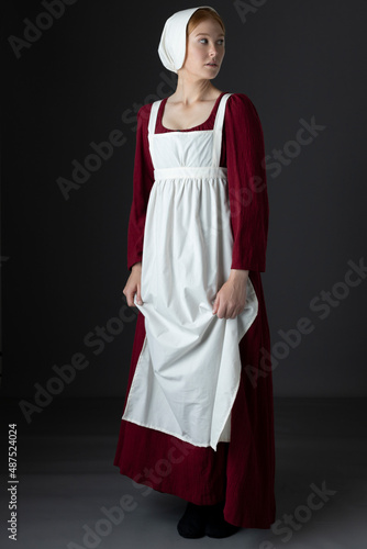 A Regency maid servant wearing a red linen dress with an apron and a cap against a studio backdrop