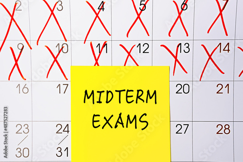 The phrase Midterm exams written on a yellow sticky note posted on a calendar or planner page as a reminder. Closeup of a personal agenda photo