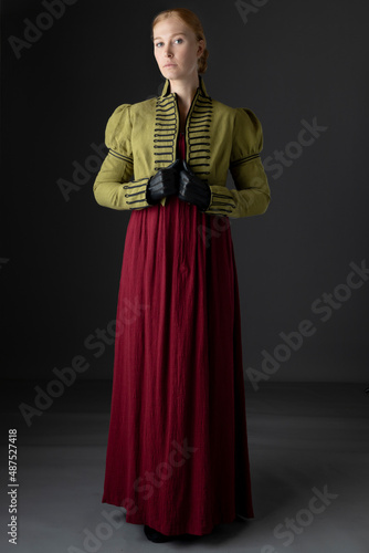 A red-haired Regency woman wearing a red linen dress with a green spencer with black braid against a studio backdrop