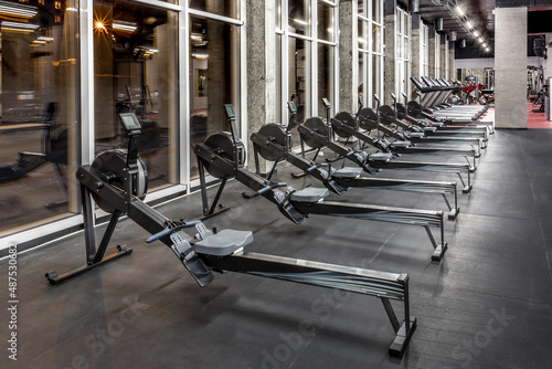Lined modern rowing machines and treadmills by window in spacious, empty gym interior. Special equipment for physical training. Sport, fitness