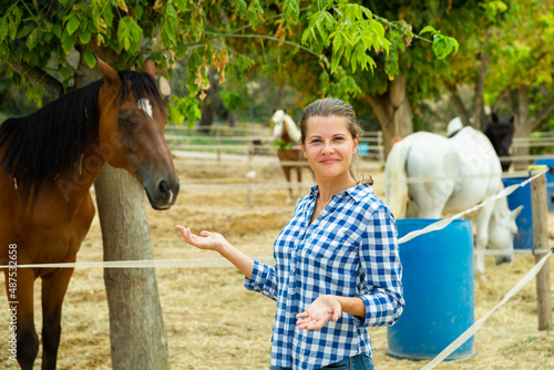 Smiling young woman standing on her ranch on background with horses in enclosure