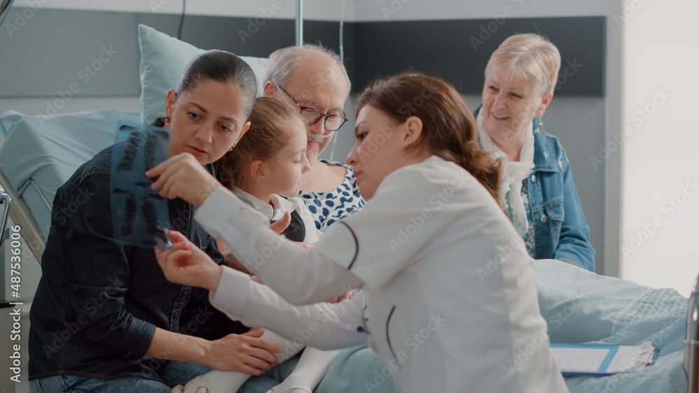 Fototapeta premium Close up of medic explaining x ray results to woman in hospital ward, talking about diagnosis of senior man. Doctor holding radiography in front of daughter, niece and wife of patient.