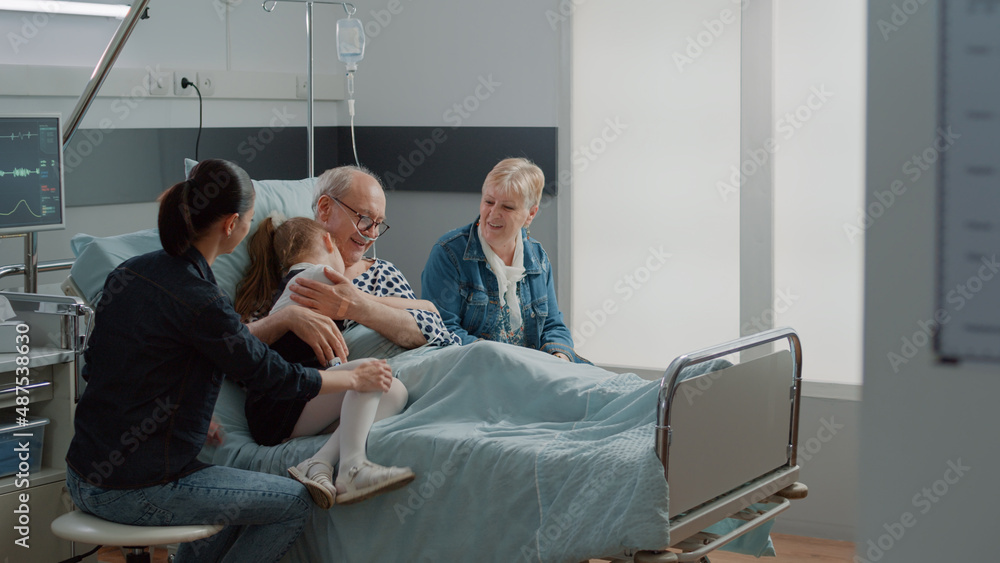 Child hugging sick grandpa in hospital ward bed at visit, comforting aged patient with illness. Kid and mother visiting senior man with IV drip bag and nasal oxygen tube. Visitors at clinic Child hugging sick grandpa in hospital ward bed at visit, comforting aged patient with illness. Kid and mother visiting senior man with IV drip bag and nasal oxygen tube. Visitors at clinic