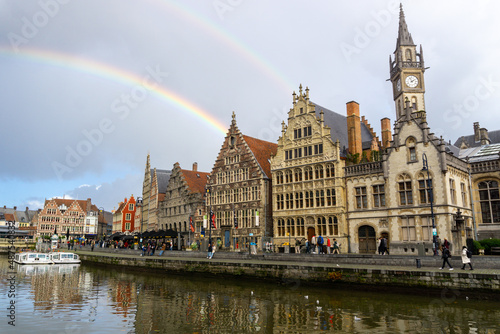 Panoramic of Ghent next to the water channels, with the reflection of the buildings in the water, with a double rainbow in the cloudy sky.