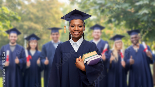 education, graduation and people concept - happy graduate student woman in mortarboard and bachelor gown with books over group of people on background photo