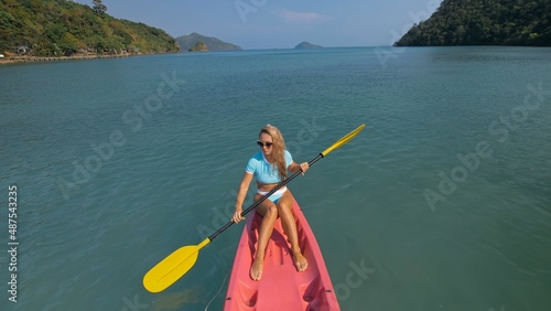 Pretty sportive blonde lady in stylish swimsuit sails on pink plastic canoe along azure ocean bay at exotic resort. Traveling to tropical countries. Girl is sailing on kayak in ocean, view from above.