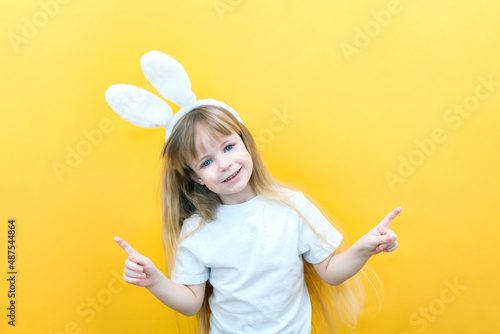 cheerful girl with rabbit ears on her head on a yellow background. Funny happy child points fingers at an empty space. copy space for text