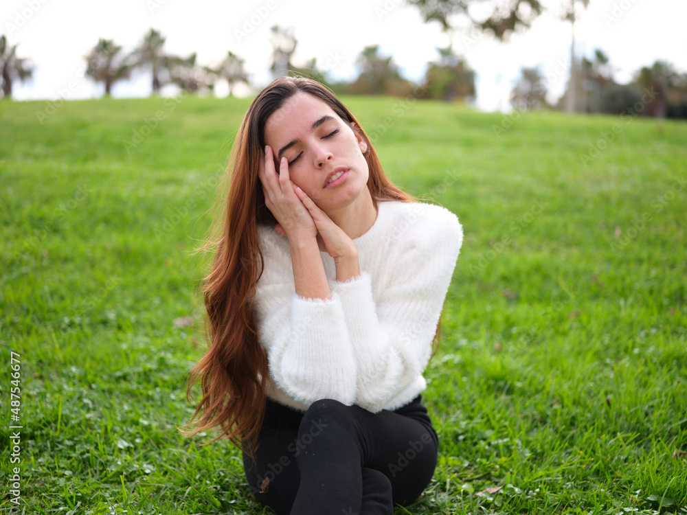 A young woman in the park sitting on the grass with her hands on her face and her eyes closed daydreaming. A young woman in the park sitting on the grass with her hands on her face and her eyes closed daydreaming.