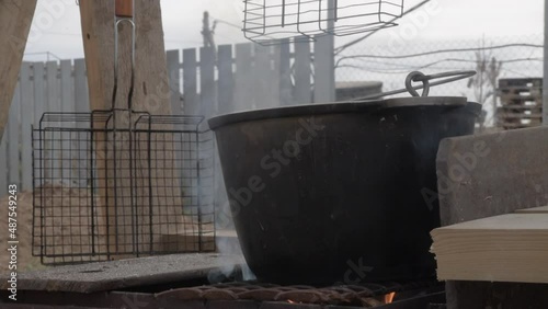 a large cauldron is on fire and there is smoke. barbecue grills hang on the background. cooking outside.