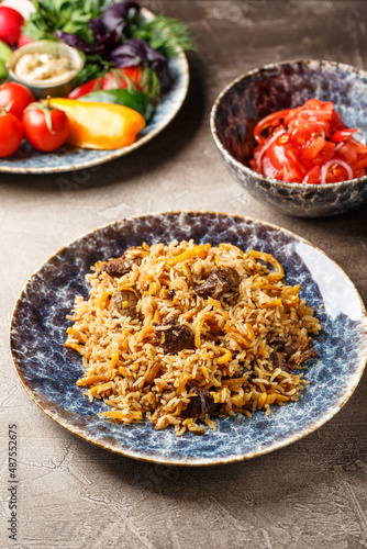 Pilaf with lamb meat in large dish, plate of tomato salad and dish with vegetables and greens
