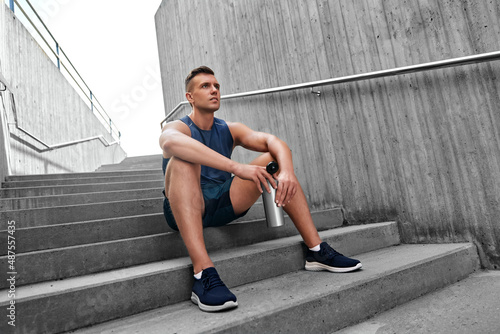fitness, sport and people concept - tired young man with bottle of water sitting on stairs