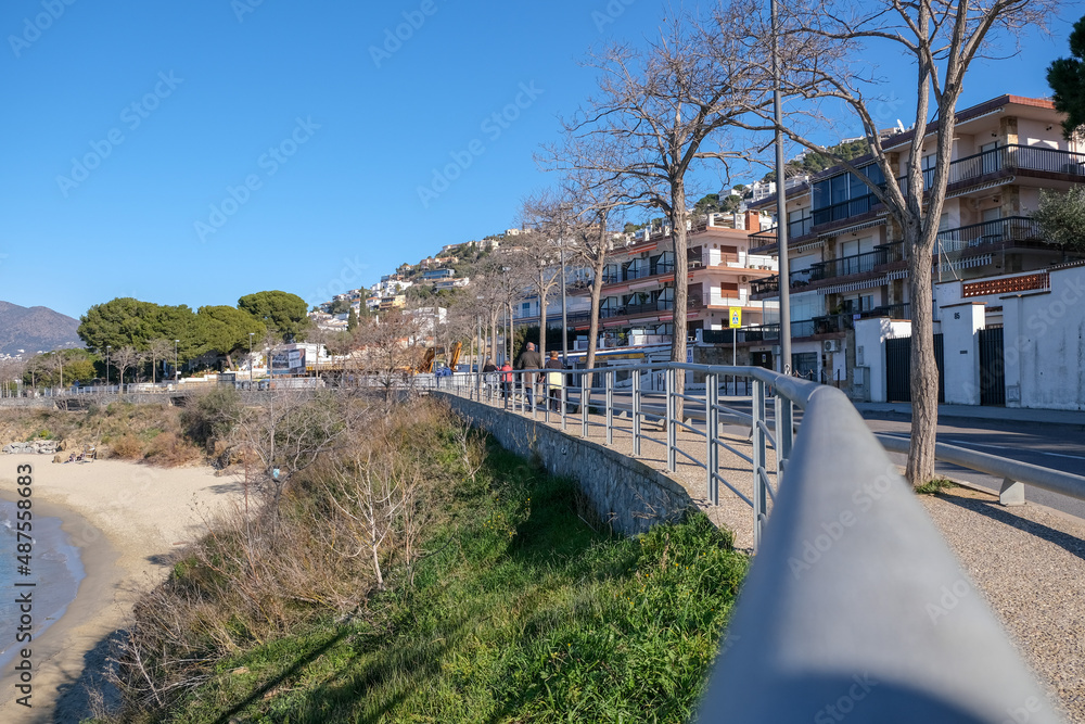 Perspective view on footpath with walking unknown people along the Mediterranean sea in Spain. Promenade seafront in Catalonia. Scenic village resort with sandy beach and traditional buildings.
