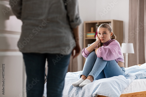 misbehavior, conflict and family concept - mother entering room and sad daughter sitting on bed at home