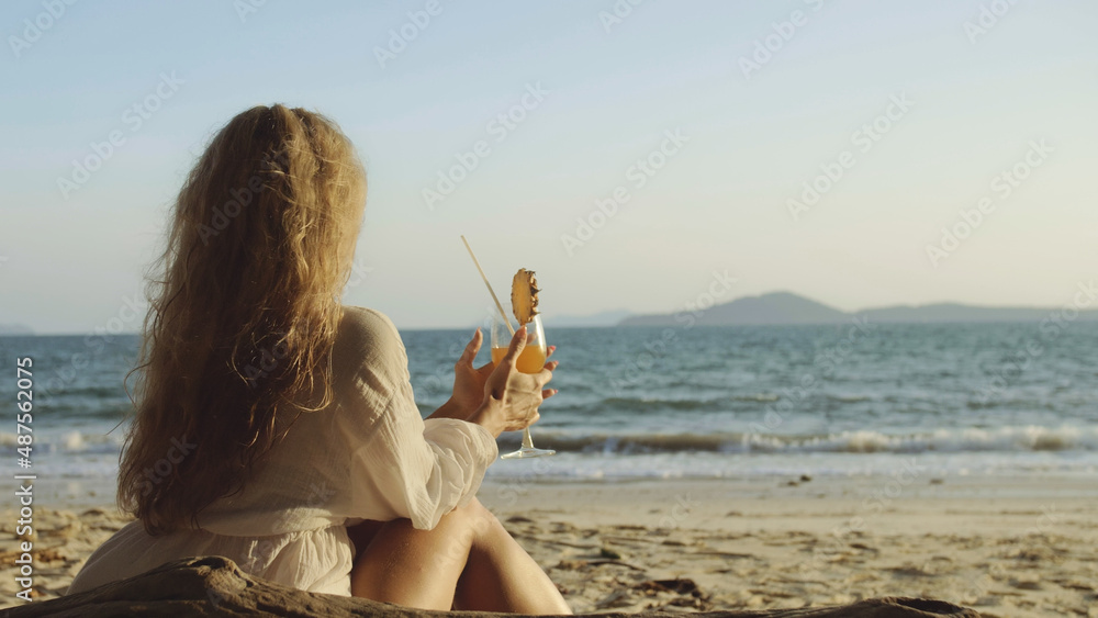 Attractive woman sitting in white dress sunglasses, drinking pineapple cocktail Pina Colada. Beautiful beach, sea waves at sunset. Concept rest tropical resort traveling tourism happy summer holidays