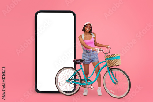 Fototapeta Naklejka Na Ścianę i Meble -  Mobile offer. Black lady riding her bike near huge smartphone with empty white screen over pink studio background