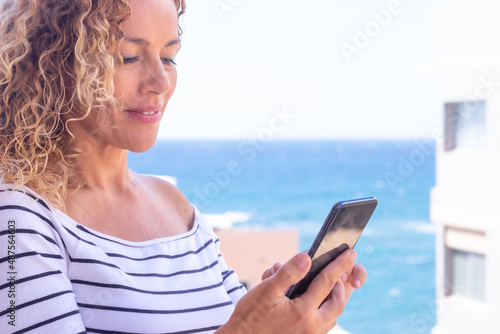 Portrait of mature blonde curly lady holding cellphone in hand standing outdoor with blue sea on background. Beautiful caucasian smiling woman using mobile phone standing on balcony.