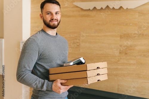 Friedly courier male holding boxes pizza and contactless payment POS terminal, standing in entrance hall of apartment or office building, by door doorway waiting to meet customer, looking at camera photo