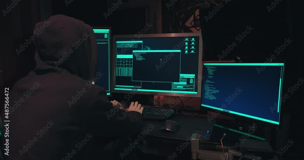 Hacker in a hood sits in front of computer screens and hacks databases. The concept of cyberterrorism, server hacking and cyberattacks. The room is dark, a person is typing code into the command line Hacker in a hood sits in front of computer screens and hacks databases. The concept of cyberterrorism, server hacking and cyberattacks. The room is dark, a person is typing code into the command line