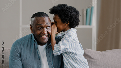 Little african american kid girl shares secret with dad tells amazing fun story in ear funny father smiling listening to cute daughter sitting on sofa trusting relationship closeness parent and child