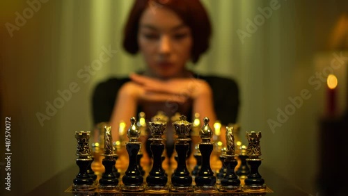 Blurred redhead female chess player moving piece thinking sitting indoors with chess board. Slim young Caucasian woman thinking strategy playing board game indoors. Concentration and lifestyle