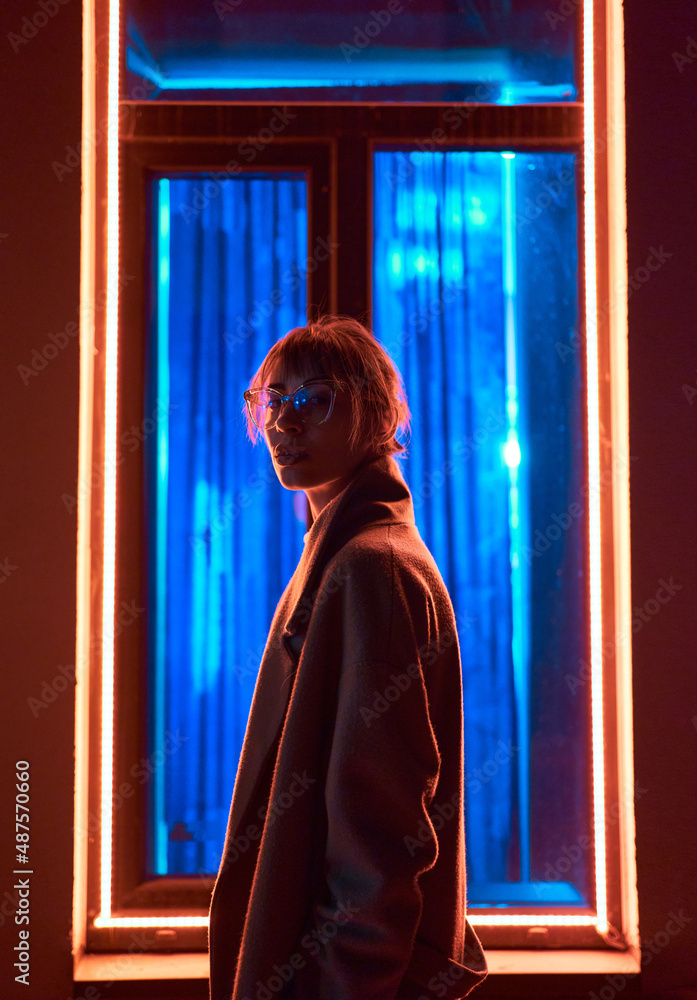 Cyberpunk girl in stylish jacket wears eyeglasses looking to camera, standing against window frame with neon lights at dark street Cyberpunk girl in stylish jacket wears eyeglasses looking to camera, standing against window frame with neon lights at dark street