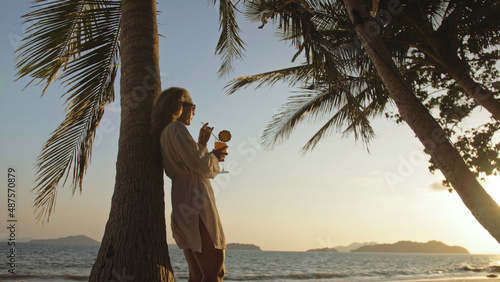 SILHOUETTE DARK GOLD SUNSET VIEW: Pretty woman drinking pineapple cocktail Pina Colada near palm tree. Woman in white shirt, sunglasses. Concept rest tropical sea traveling tourism summer holidays