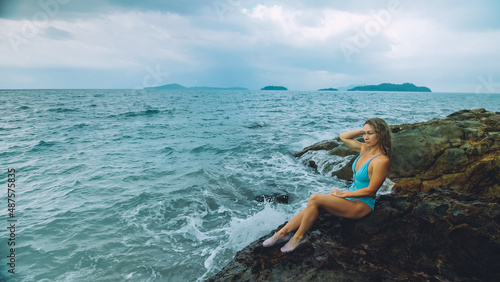 Alone attractive woman walk near a rock reef hill in stormy morning rain cloudy sea. Girl in turquoise swimsuit. Concept resort outdoor relax, vacation, loneliness. Dark dramatic view