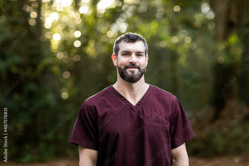 A doctor with dark hair and a beard in maroon scrubs standing outside in a natural green environment