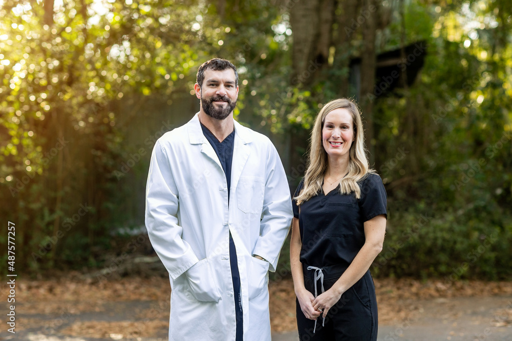 A doctor with dark hair and a beard in black scrubs and a white lab coat standing outside in a natural green environment talking to another medical professional A doctor with dark hair and a beard in black scrubs and a white lab coat standing outside in a natural green environment talking to another medical professional