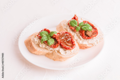 Bruschettas or toasts with ricotta cheese and sliced sun dried tomatoes topped with basil on white plate, white table background. Delicious italian appetizer, antipasti