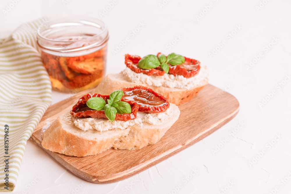 Delicious toasts with ricotta cheese, sliced sun dried tomatoes topped with basil and dried tomatoes with olive olive in jar on wooden serving board, white table background. Classic appetizer