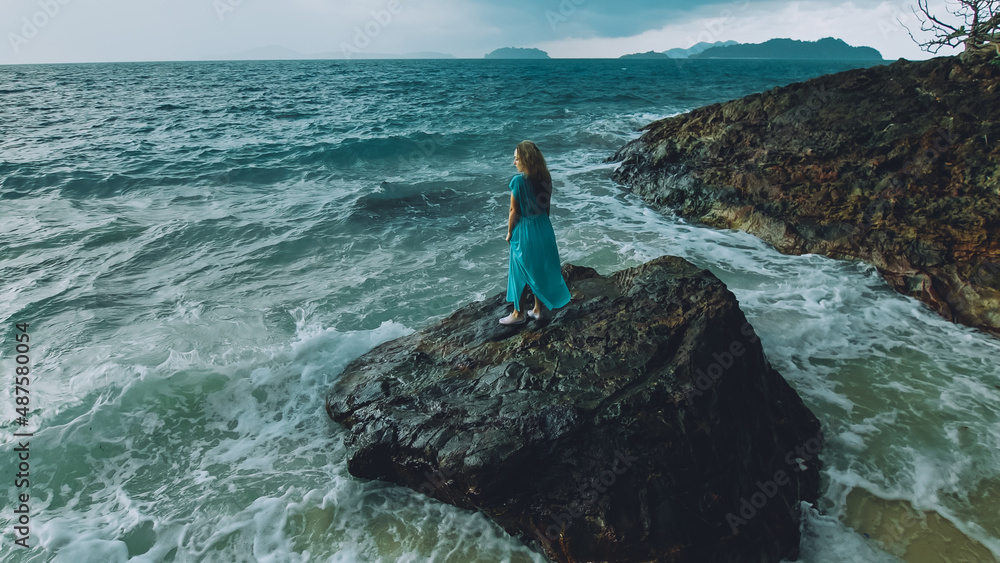Sexy hot woman meditates, relaxes on a rock reef hill in stormy morning rain cloudy sea. Girl in blue swimsuit, dress tunic. Concept feminine, relax, sexual health. Dark dramatic silhouette view