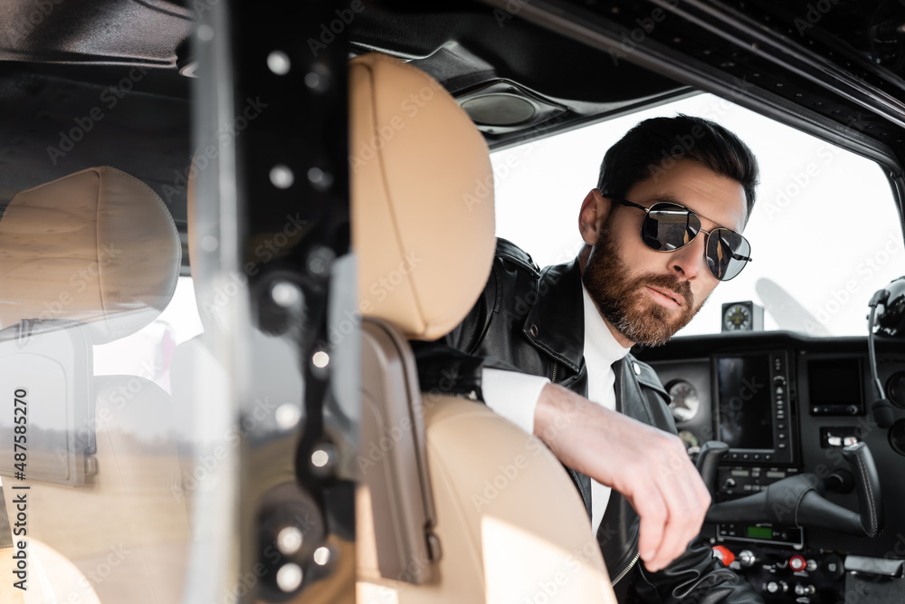 bearded pilot in stylish sunglasses and leather jacket sitting in cockpit of helicopter.